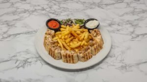 Sliced chicken shawarma arranged around golden fries, served with garlic sauce, tomato sauce, and fresh onion and herb salad on a white plate.