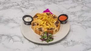 Sujuk shawarma slices arranged around seasoned fries, served with creamy garlic sauce, tomato sauce, and a fresh herb salad on a white plate.
