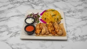 Grilled chicken fillet skewers served with yellow basmati rice, fresh onion salad, flatbread, yoghurt dip and red sauce on a white plate.
