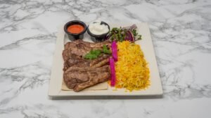 Grilled lamb chops served with yellow basmati rice, fresh salad, pickled onions, and dipping sauces on a white plate. Copy alt text